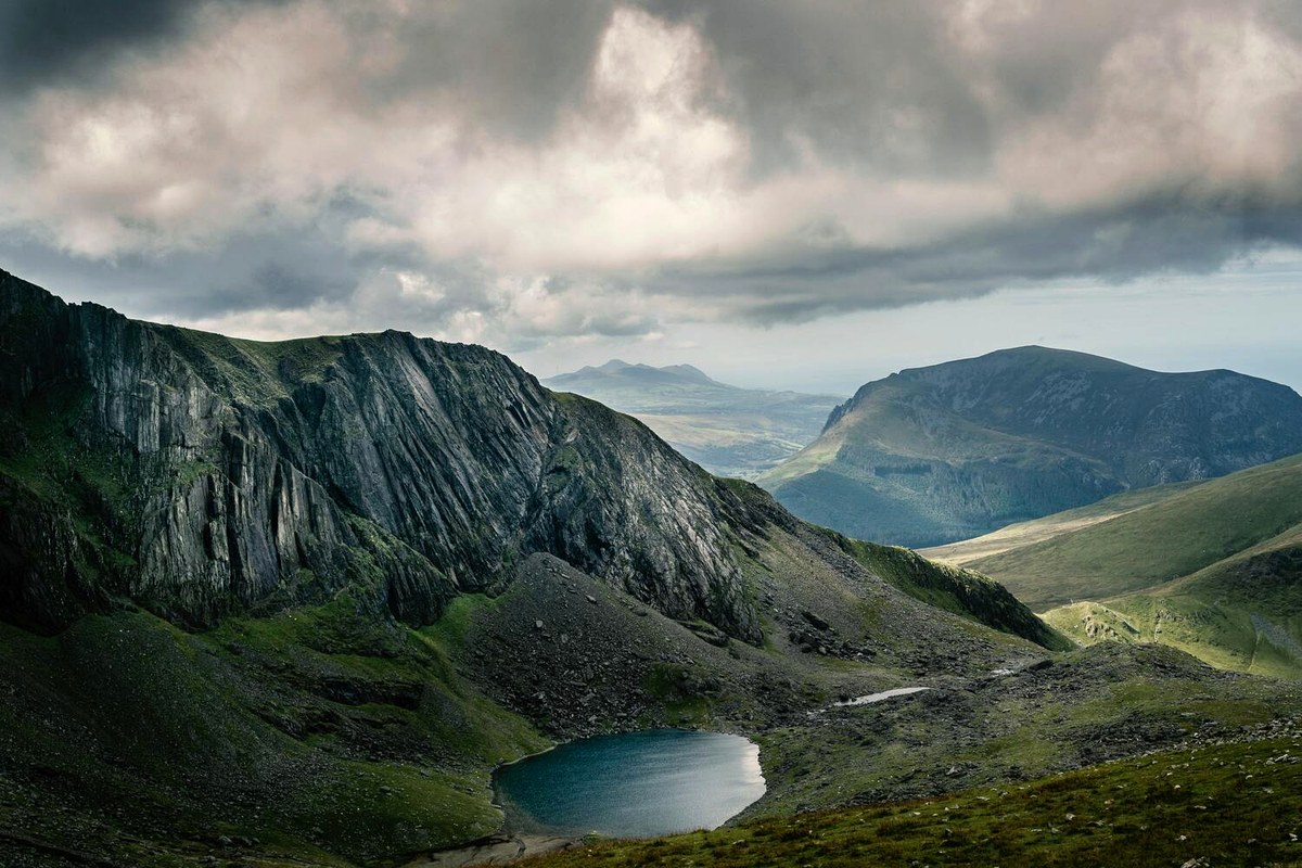 A dramatic mountain valley with layered ridges, a small lake, and moving cloud.